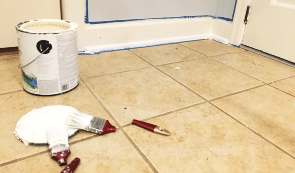 two paintbrushes against a tan tile floor, background includes wall with painters tape outline a freshly painted triam and molding.